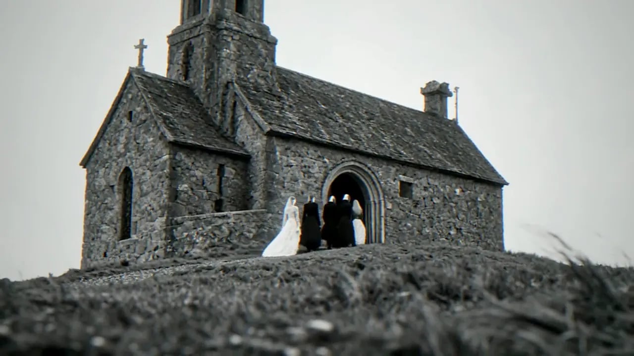 A bride in a white veil and gown stands with nuns at the arched door of a stone chapel on a grassy hilltop, at 0:56 in Woke Up Dead